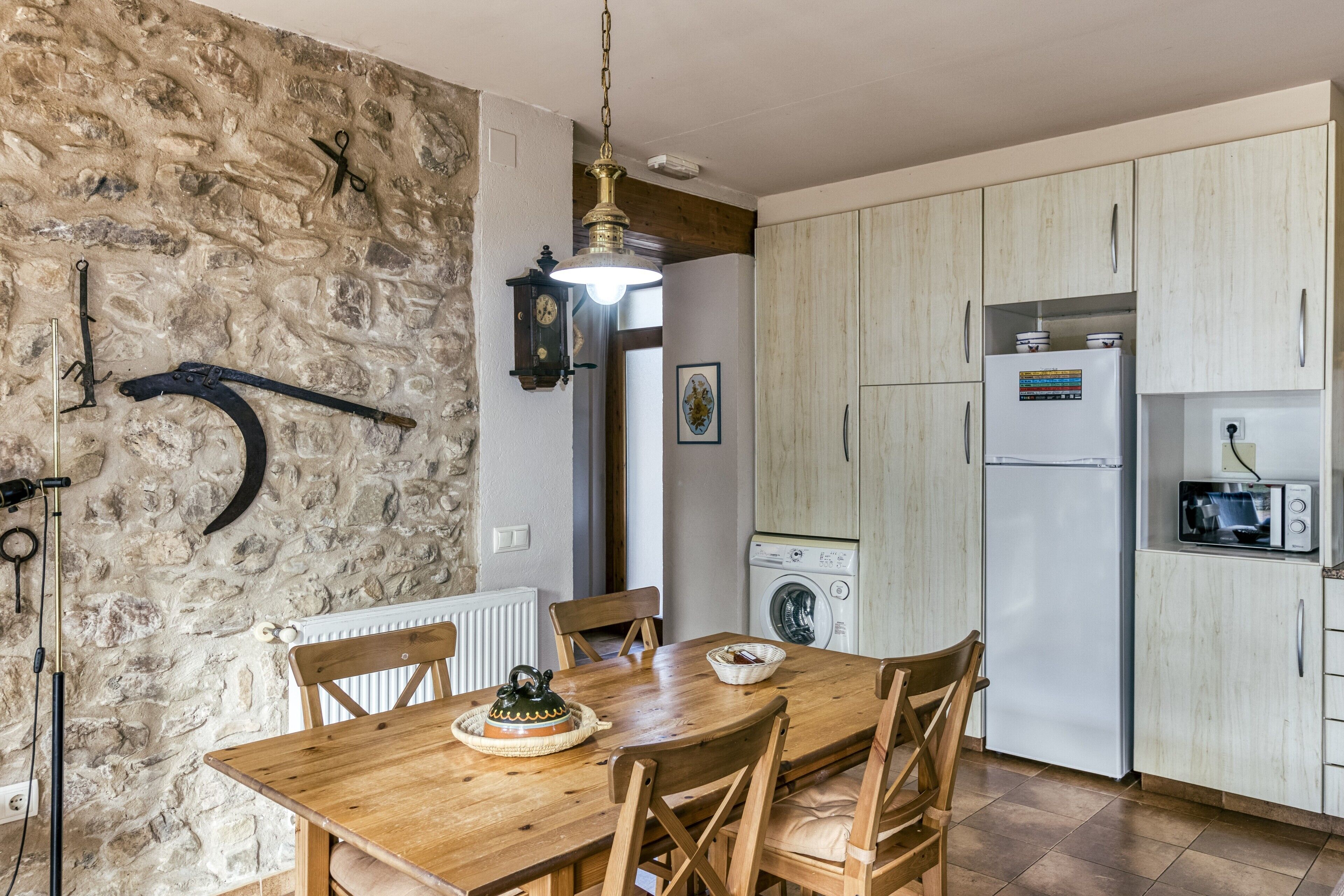 Una cocina con pared de piedra, mesa de madera, sillas, lavadora y refrigerador.
