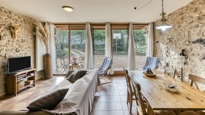 Una sala de estar acogedora con sofá, televisión, mesa de comedor y sillas, ventanas amplias con cortinas y una pared de piedra.