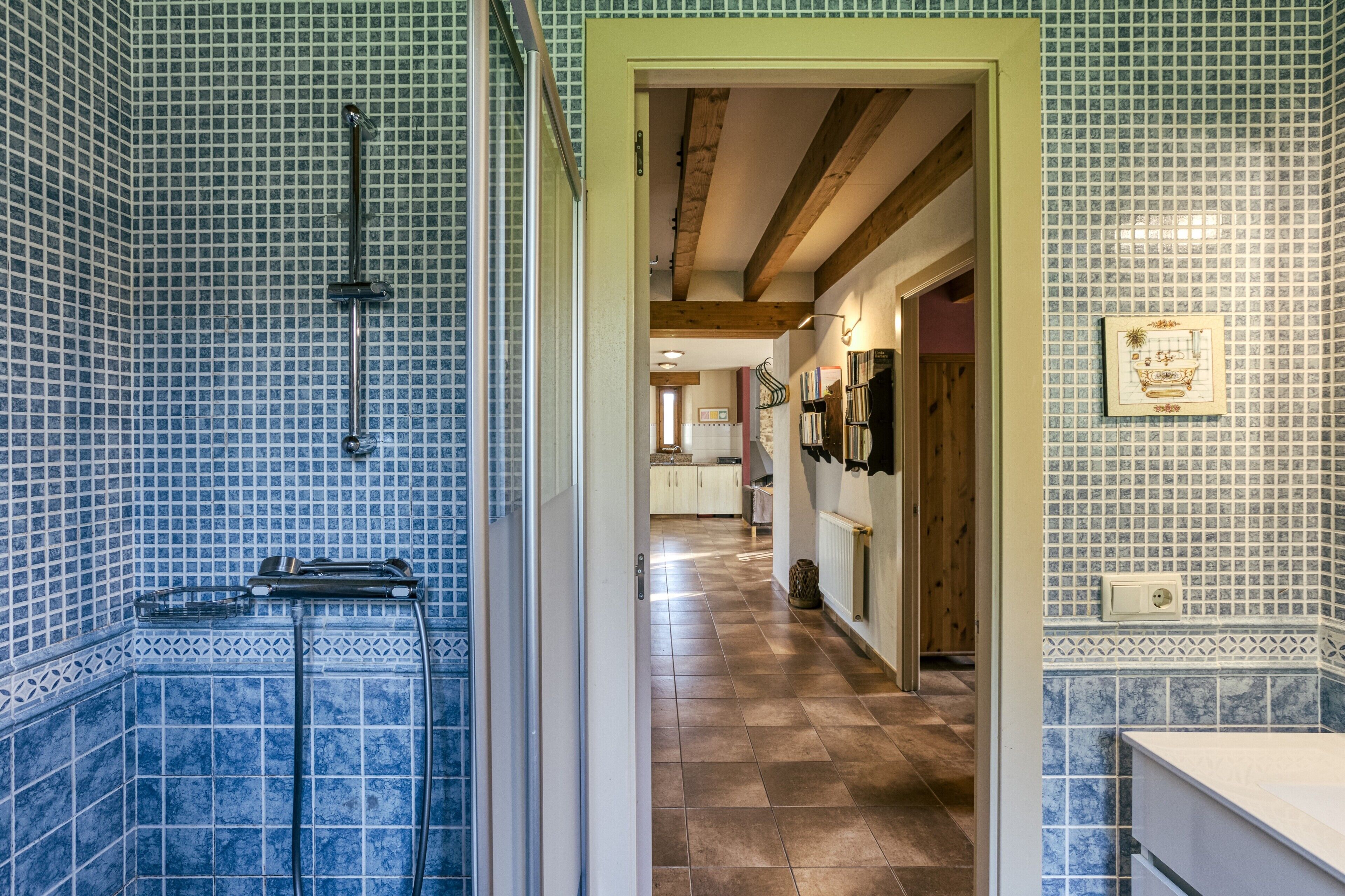 Baño con paredes de azulejos azules, zona de ducha y pasillo que conduce a una cocina.