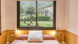 Une chambre avec une grande fenêtre, un lit agrémenté d’un coussin à motifs, des lampes de chevet et une vue sur un jardin et un bâtiment.