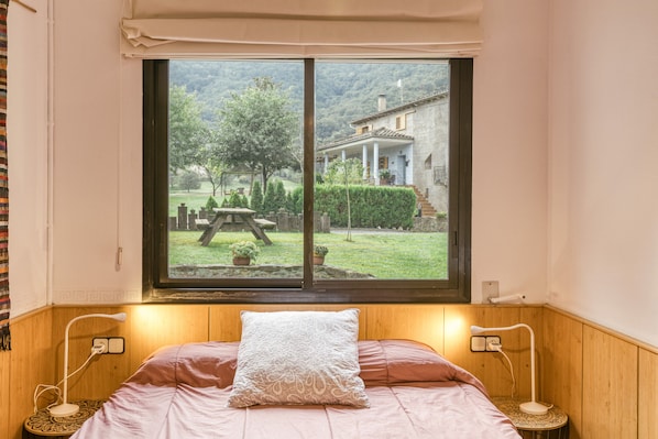 Un dormitorio con un ventanal grande, una cama con almohada estampada, lámparas de noche y vistas a un jardín y un edificio.