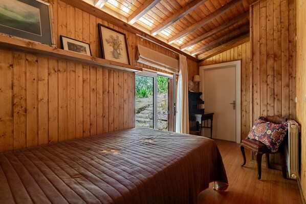 A wooden room with a bed, a chair, a radiator, and a door leading outside.