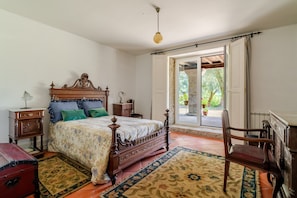 Ein Schlafzimmer mit einem hölzernen Bett, einem Nachttisch, einem Sessel, einer Kommode und einem Fenster mit Blick auf Grünflächen.
