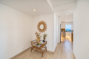 Ein modernes Interieur mit Essbereich, ausgestattet mit einem Glastisch, einem dekorativen Spiegel und einem Korbstuhl. Im Hintergrund ist eine Küche mit Meerblick zu sehen.