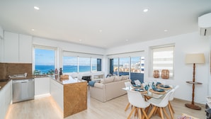 Une cuisine moderne équipée d’un lave-vaisselle, un coin repas avec une table et des chaises en verre, un canapé et de grandes fenêtres offrant une vue sur la mer.