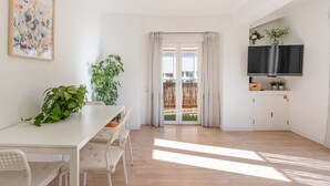 Quarto moderno com uma mesa branca, cadeiras, uma TV e uma varanda com cortinas.