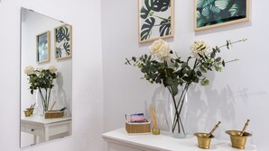 Uma toucador branca com espelho, quadros enquadrados, um vaso com flores e uma pequena cesta com revista e incensos.