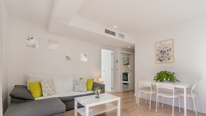 Uma sala de estar moderna com sofá cinzento, mesa de centro branca e uma área de jantar com cadeiras e mesa brancas.