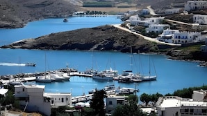 Uma marina costeira com barcos atracados, cercada por edifícios brancos com detalhes azuis, com colinas ao fundo.