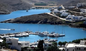 A coastal marina with boats docked, surrounded by white buildings with blue accents, set against a backdrop of hills.