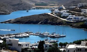 A coastal marina with boats docked, surrounded by white buildings with blue accents, set against a backdrop of hills.