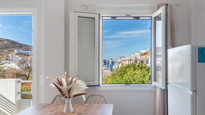 Una habitación con una mesa de madera, un florero con plantas secas, una ventana que ofrece vistas abiertas a edificios y follaje, y un refrigerador blanco.