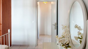 Un pasillo con una puerta blanca, un marco de madera, piso de baldosas blancas y un espejo con un arreglo floral blanco.
