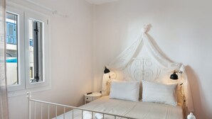 Un dormitorio con cama blanca, mesita de noche, ventana y cortina.