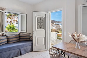 A room with a sofa, a dining table, and a door leading to a balcony with a view of buildings and trees.