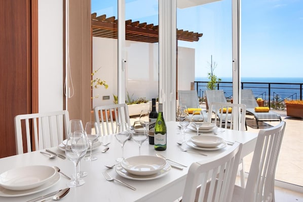 A dining table set for four with white chairs, a bottle of wine, and a view of the sea.