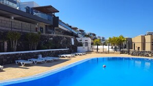 A swimming pool with lounge chairs, a stone wall, and modern buildings in the background. - Holiday Apartment 'Obsidian Villa Bentayga' with Sea View and Wi-Fi (Adeje)