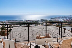 A balcony with wicker chairs, a white planter with plants, and a view of the sea and cityscape.