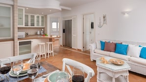 A modern kitchen with white cabinets, a dining area with a table set for four, and a living room with a sofa and cushions.
