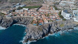 An aerial view of a coastal town with a rocky shoreline, residential buildings, and a large hotel with a pool.