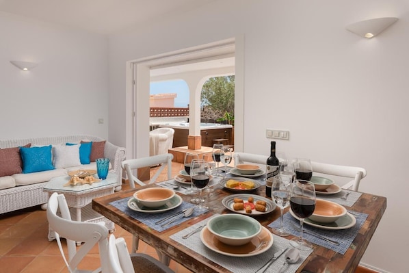 A dining area with a table set for four, surrounded by white chairs, with a view of a patio and outdoor furniture.