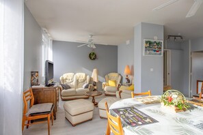 Una sala de estar con ventilador de techo, una mesa de comedor con un centro de mesa floral y un televisor sobre un mueble bajo.