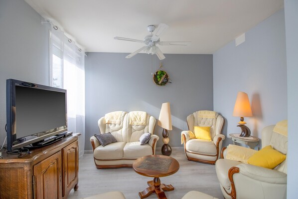 A living room with a ceiling fan, a flat-screen TV, two armchairs, a small round table, and a lamp. - Bright Apartment with Garden in the Heart of Le Bugue (Le Bugue)