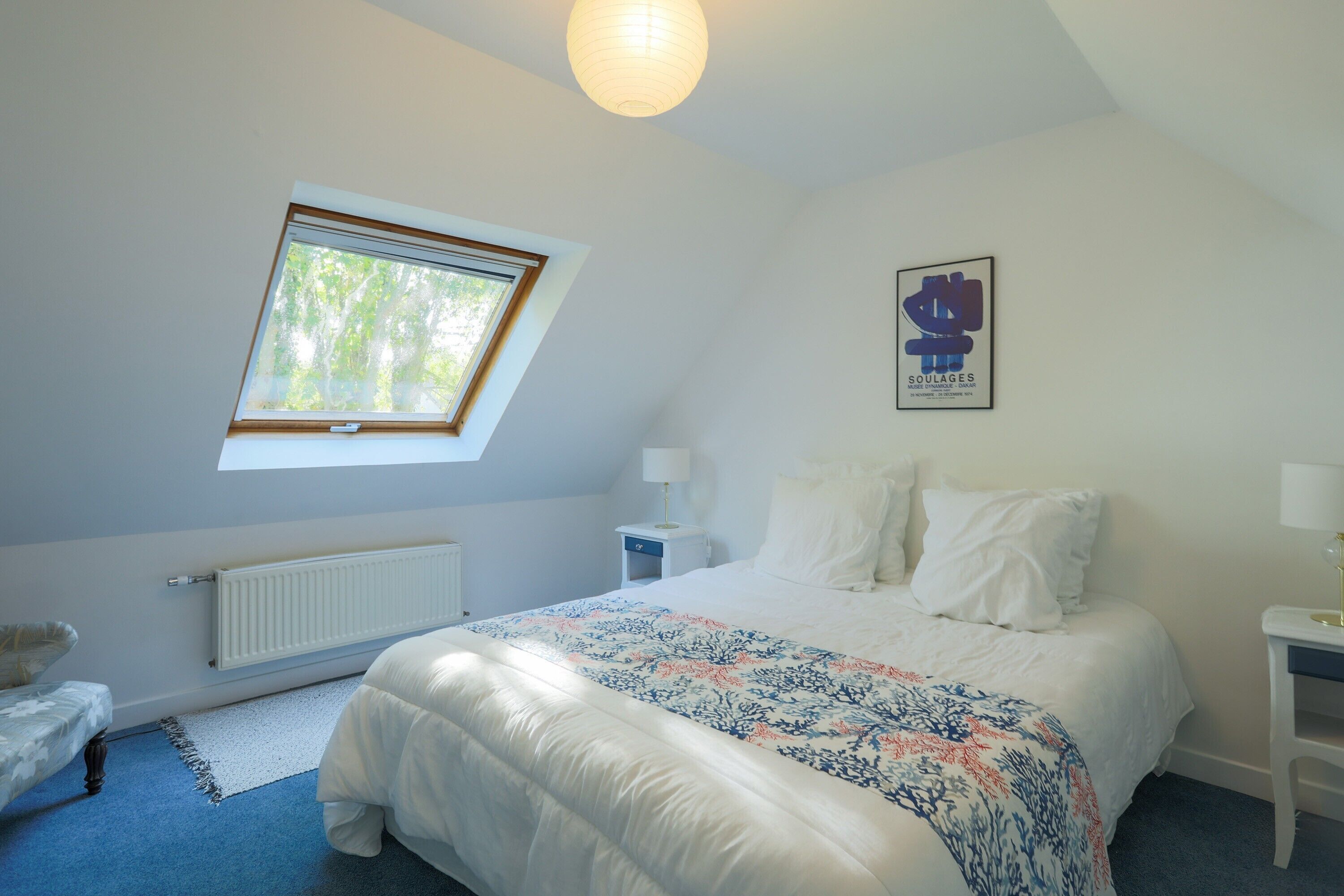 A bedroom with a bed, a window, a radiator, a nightstand, a lamp, and a framed picture on the wall.