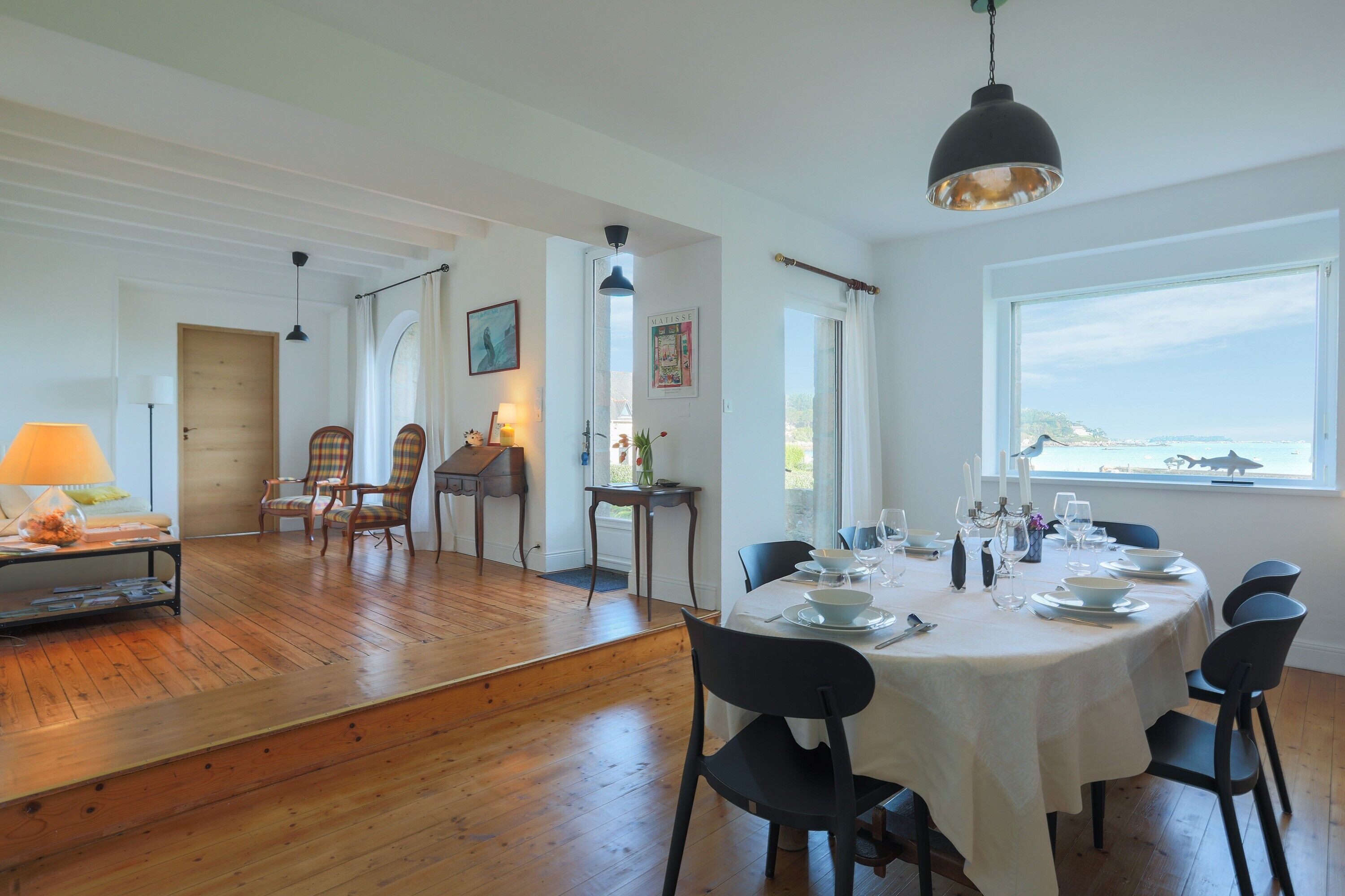 Un coin repas avec une table dressée pour quatre personnes, un plancher en bois et une vue sur la mer par une grande fenêtre.