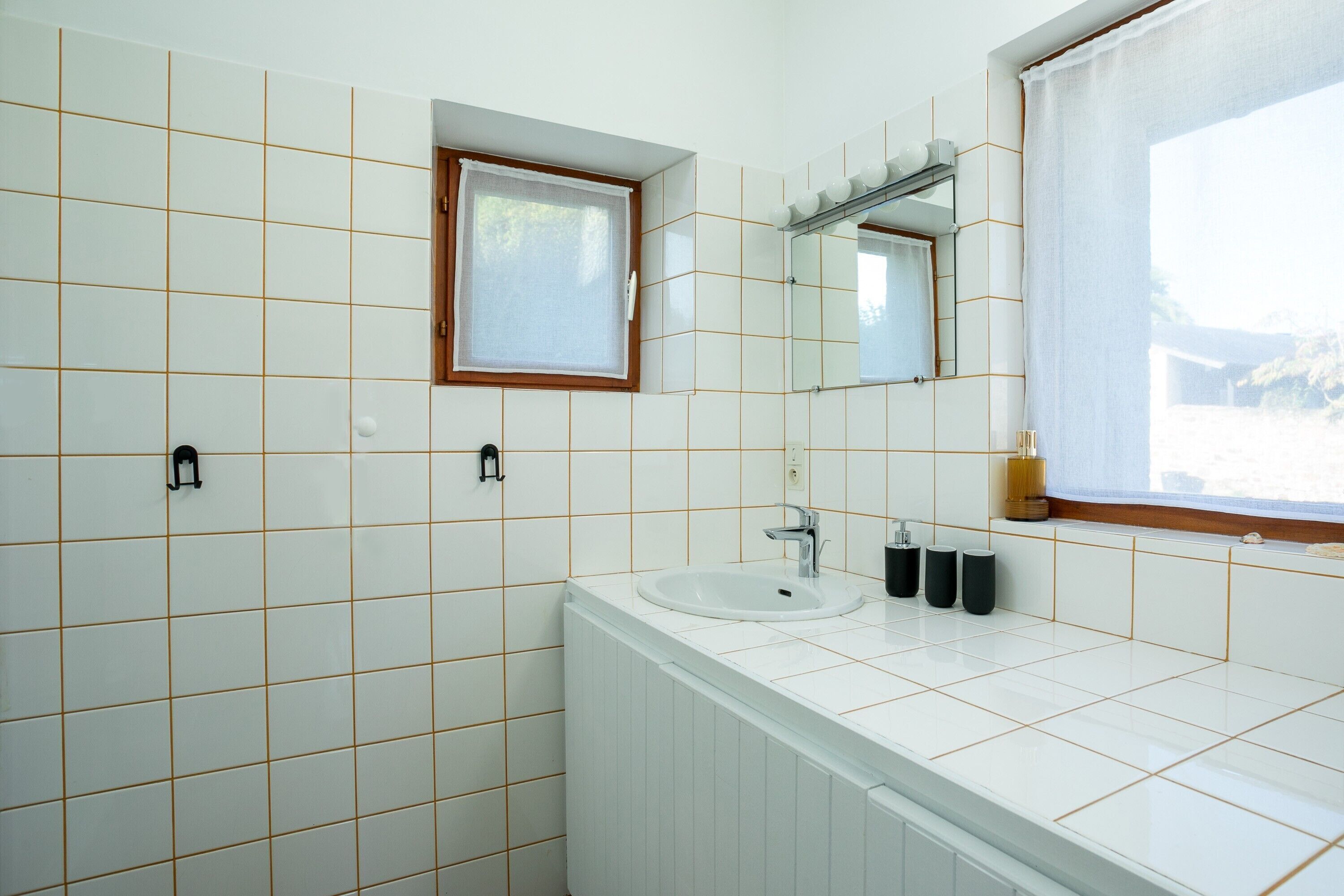 A bathroom with white tiled walls, a window with a wooden frame, a sink, a mirror, and a radiator.
