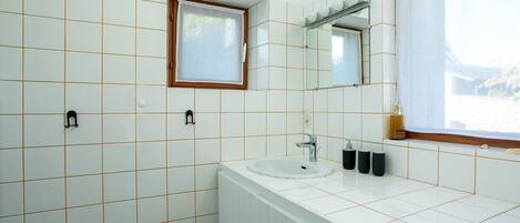 A bathroom with white tiled walls, a window with a wooden frame, a sink, a mirror, and a radiator.