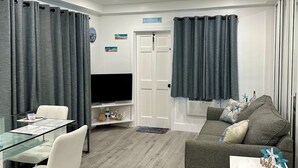 A modern living room with a glass dining table, white chairs, a grey sofa, and a flat-screen TV. - Newly renovated apartment with ocean views, steps from the beach and Boardwalk! (Hollywood)