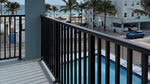 A balcony with a view of a swimming pool, palm trees, and a building with multiple windows. - Newly renovated apartment with ocean views, steps from the beach and Boardwalk! (Hollywood)