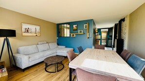 A living room with a sofa, a coffee table, a dining table with chairs, a lamp, and a kitchen area visible through an open door.