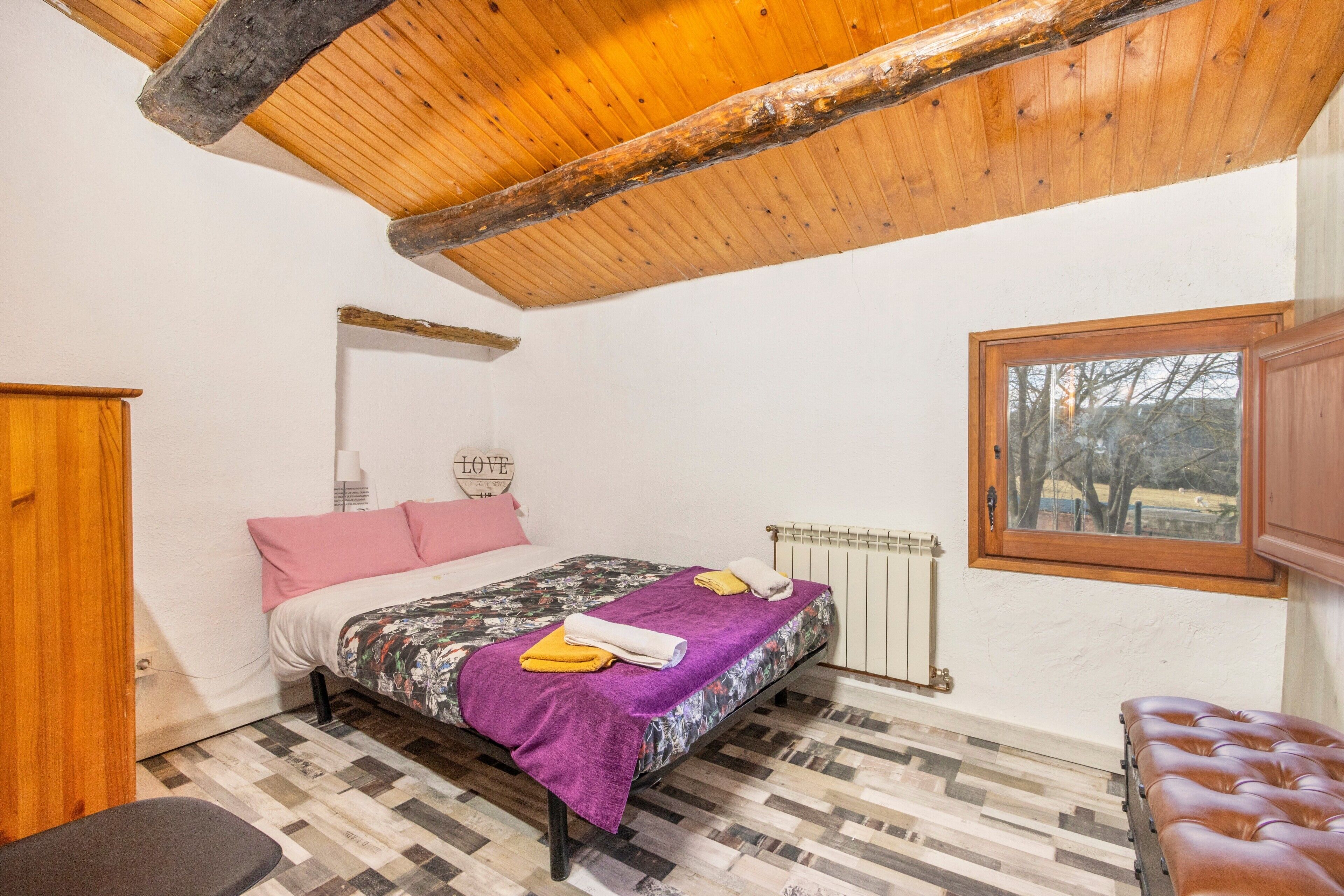 A bedroom with a wooden ceiling, a bed with a patterned bedspread, a radiator, and a window with a view.