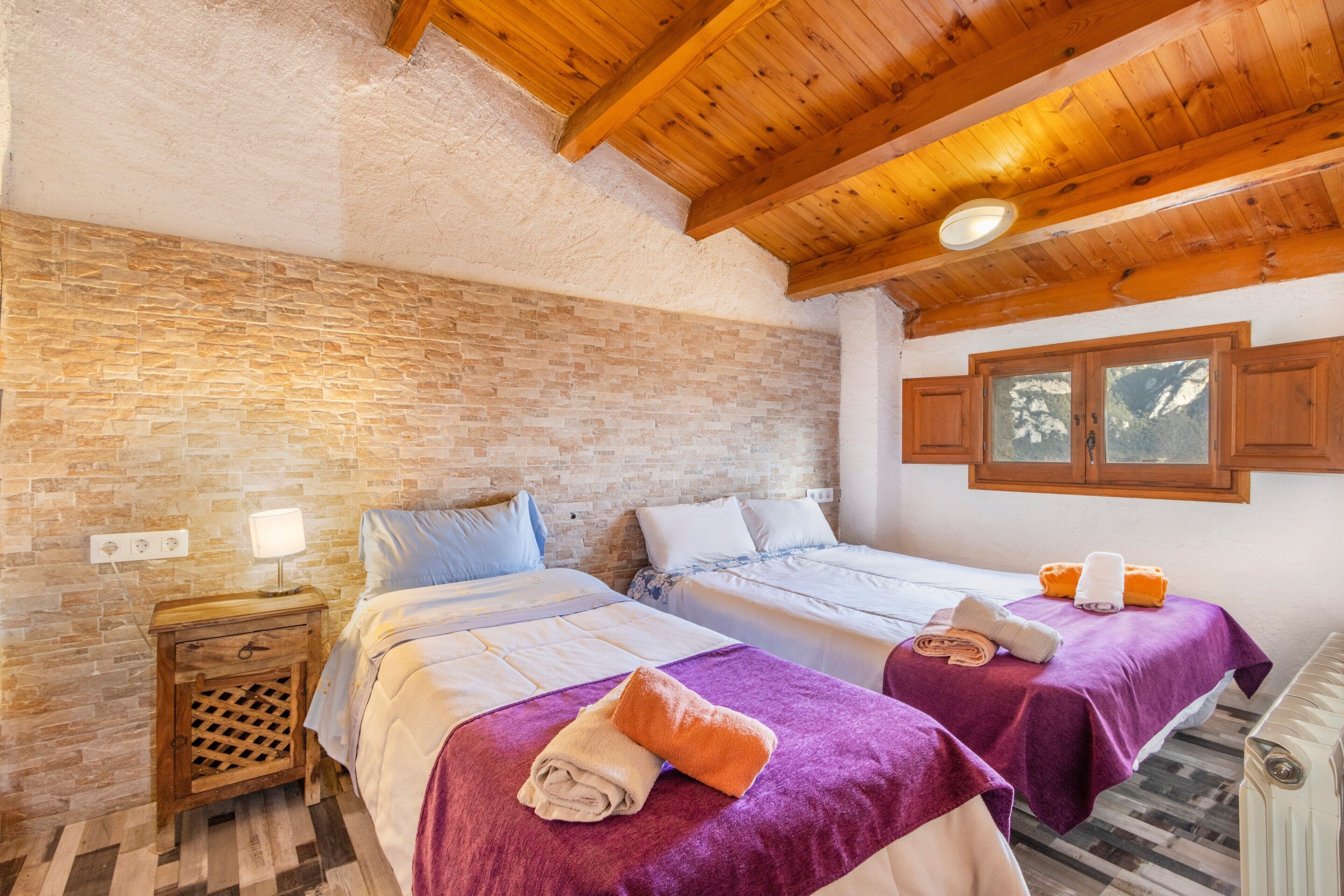 A room with two beds, wooden ceiling beams, a brick wall, and a window with a view.