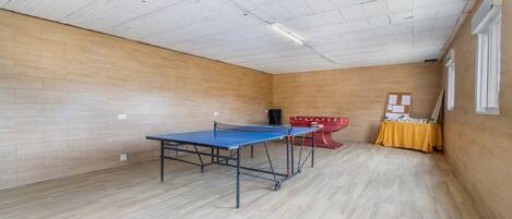 A room with a ping pong table, a red table tennis table, and a yellow table with a sign.