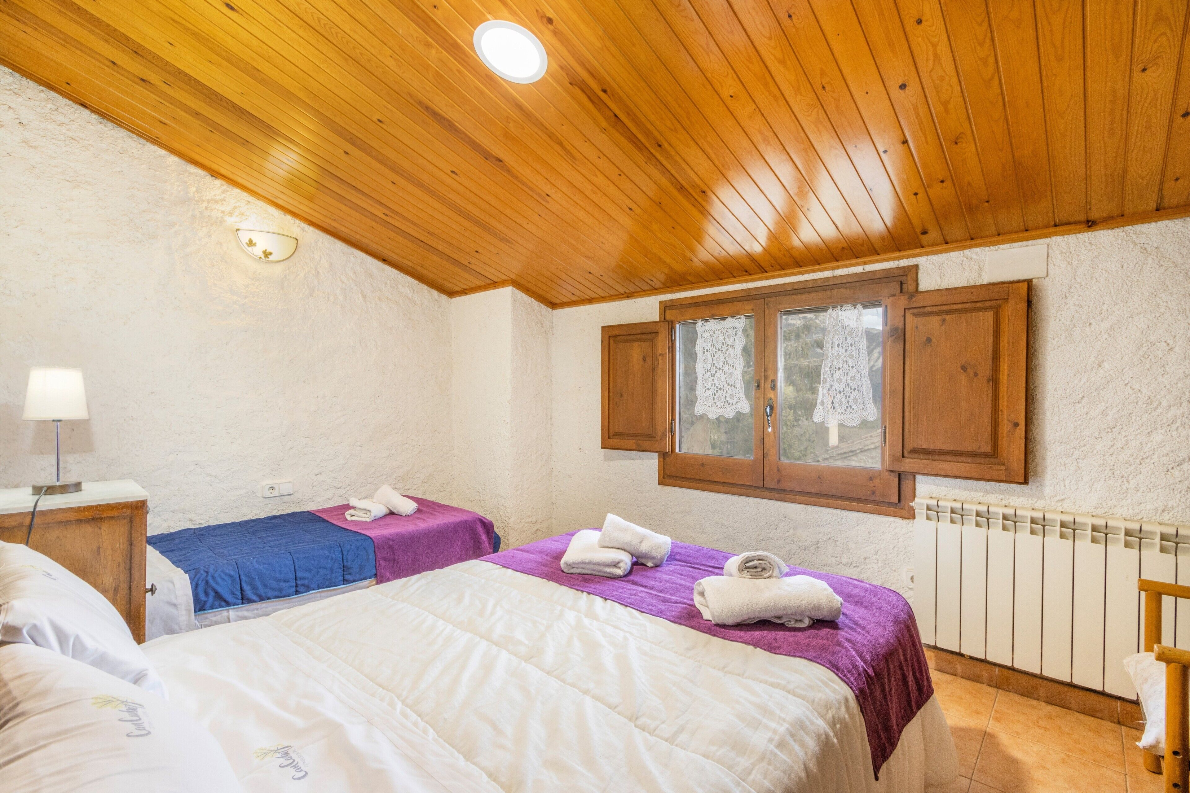 Een slaapkamer met twee bedden, een houten plafond en een raam met houten luiken.
