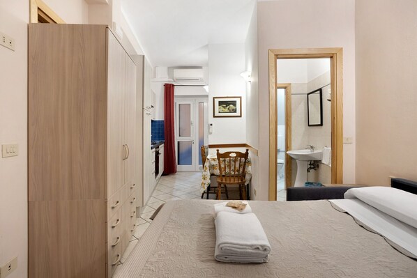 A hotel room with a bed, a desk, a chair, a wardrobe, a bathroom with a sink, and a mirror.