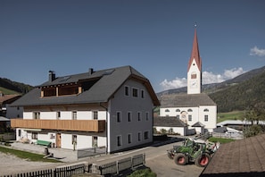 Um edifício de dois andares com varanda, uma igreja com uma torre alta e um trator em primeiro plano.