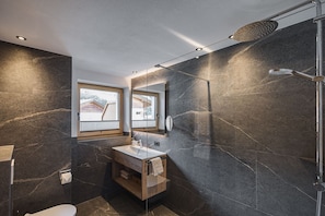 A modern bathroom with a large mirror, a wooden vanity, a shower, and a view of a house through a window.