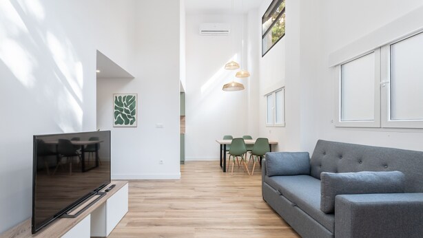 Una sala de estar moderna con un televisor de pantalla plana, un sofá gris, una mesa de comedor con sillas, lámparas colgantes y un ventanal grande.