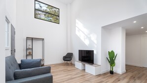 Una sala de estar moderna con un sofá gris, una silla negra, un televisor de pantalla plana sobre un soporte blanco, un ventanal con vistas a árboles y una planta en maceta.