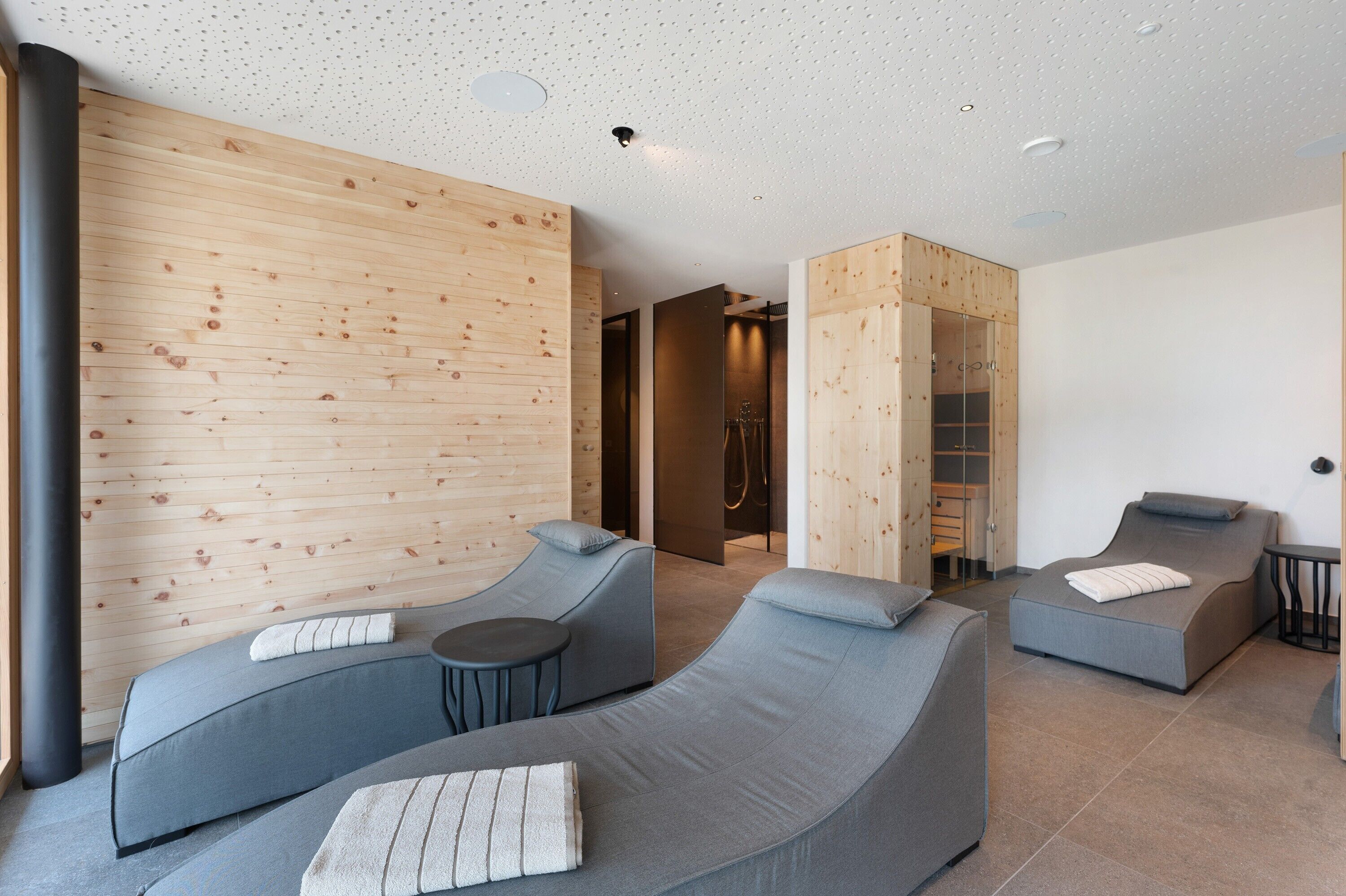 Ein moderner Wellnessraum mit Holzpaneelen, zwei Liegemassagesesseln, einem kleinen Tisch und einer begehbaren Dusche.