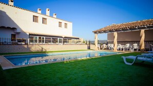 Una piscina con una zona verde, un edificio bianco con finestre e un portico coperto con sedie.