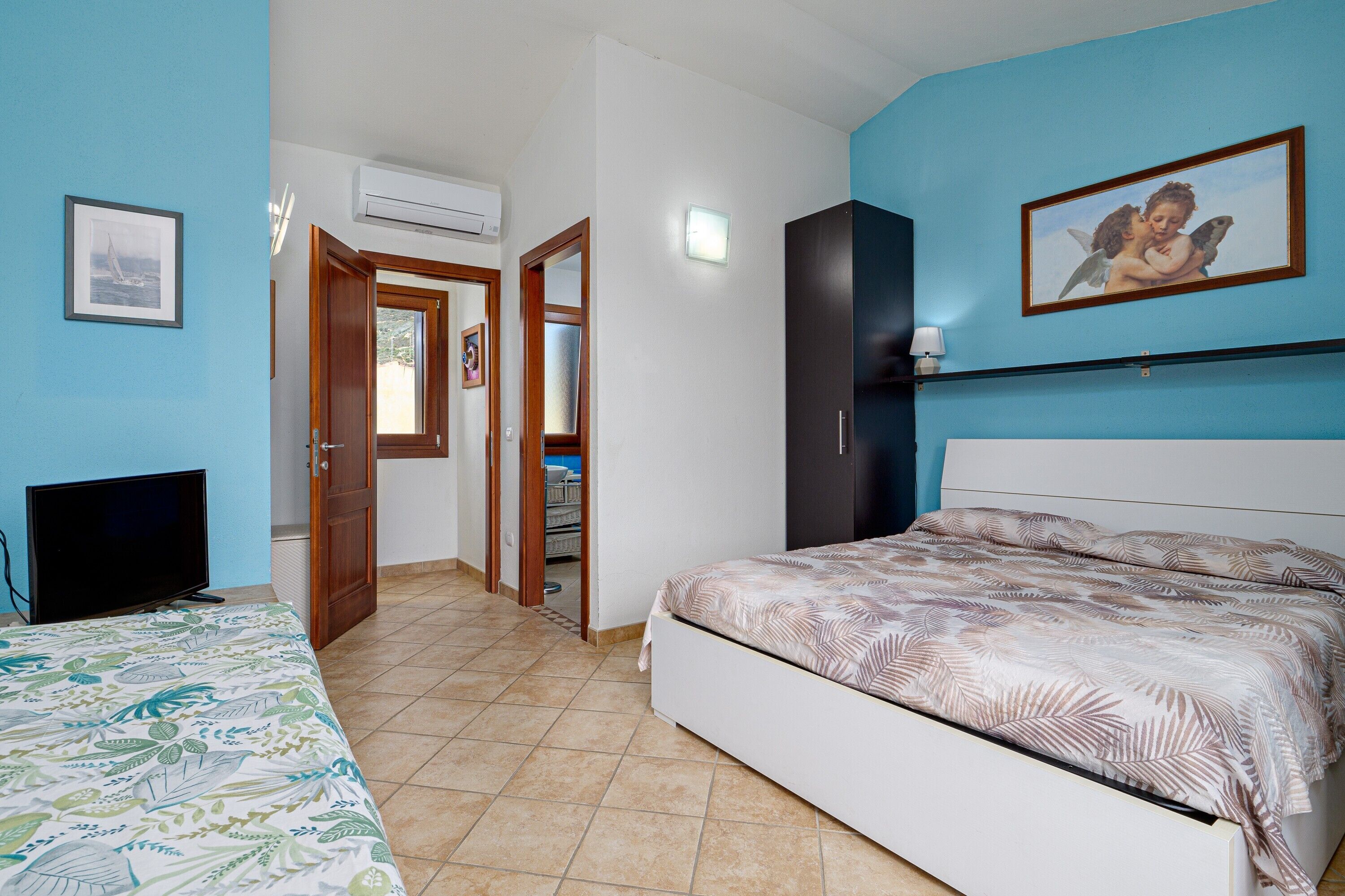 A bedroom with a bed, a television, a door leading to another room, and a wall-mounted air conditioner.