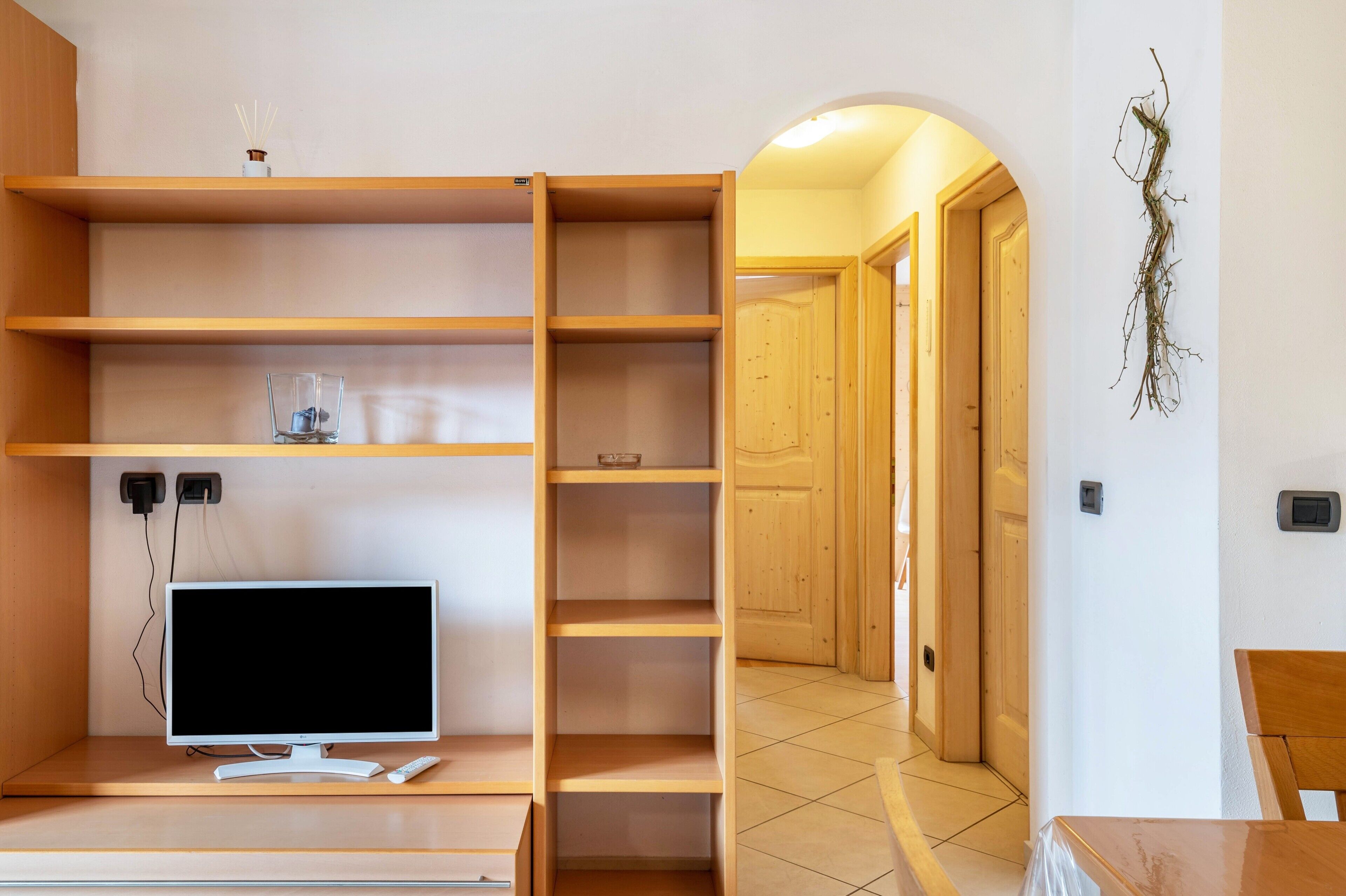 Una habitación con una televisión sobre un mueble bajo, estantes de madera, una mesita y una puerta que da a otra estancia.