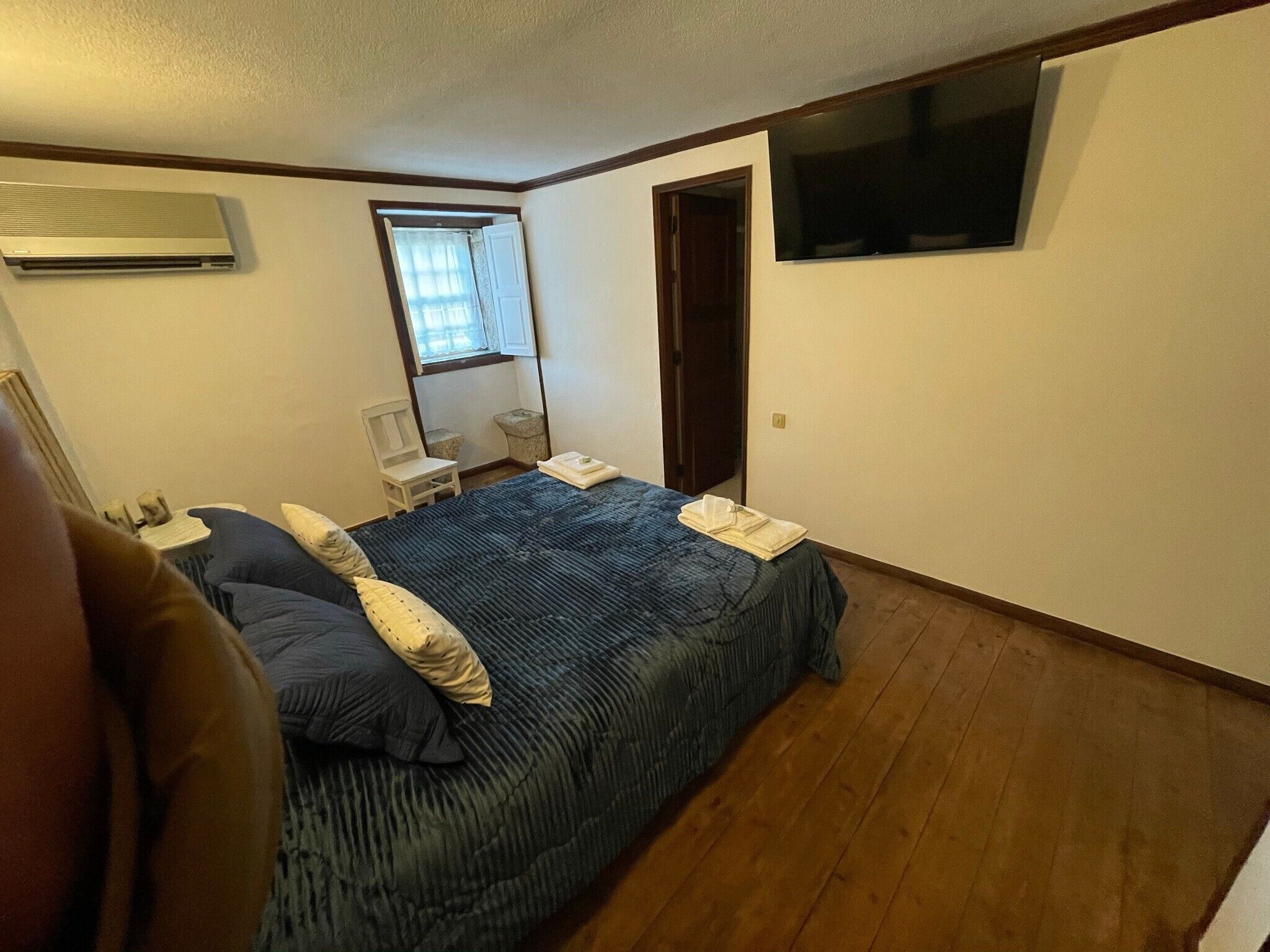 A bedroom with a bed, a chair, a desk, a window, and a TV mounted on the wall.