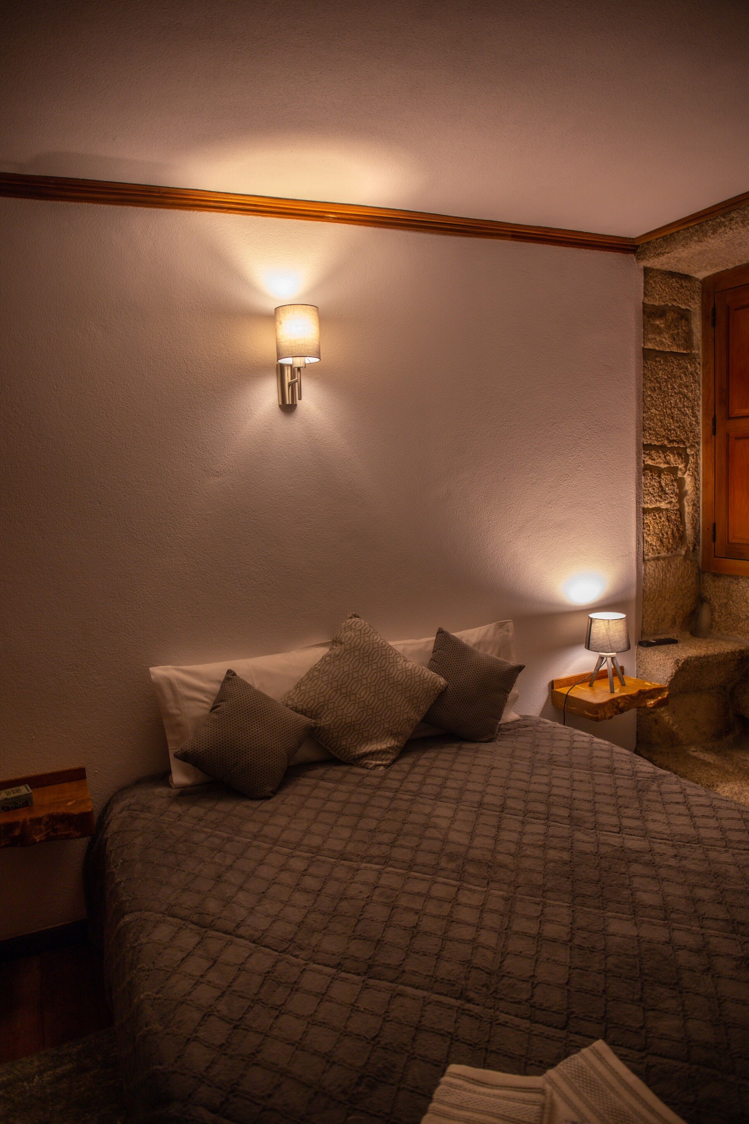Une chambre à coucher comprenant un lit, des oreillers, une table de chevet, une lampe fixée au mur et un mur d’accent en pierre.