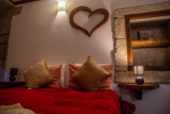 A bed with red bedding, a heart-shaped wall decoration, a shelf with a book, and a lamp.